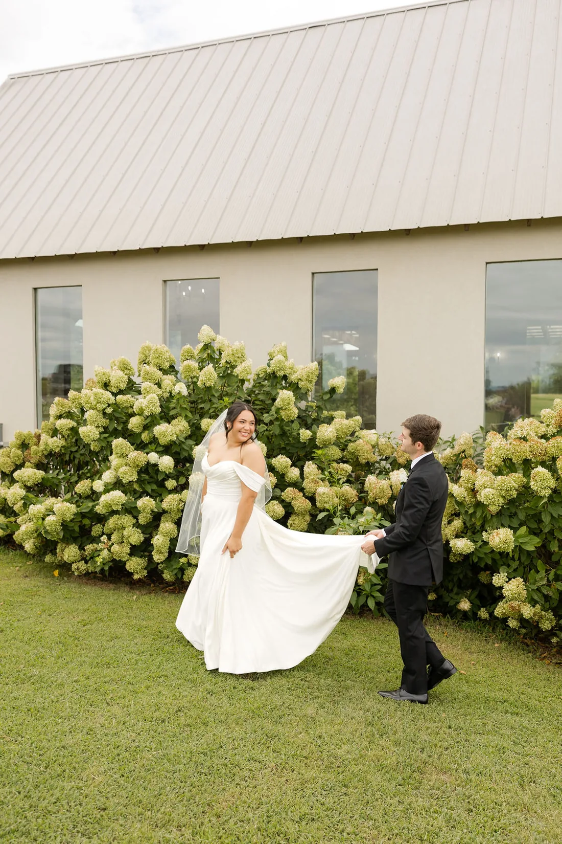 Jennifer by the white hydrangeas as Hayden holds her train at Gateley Hill Gardens
